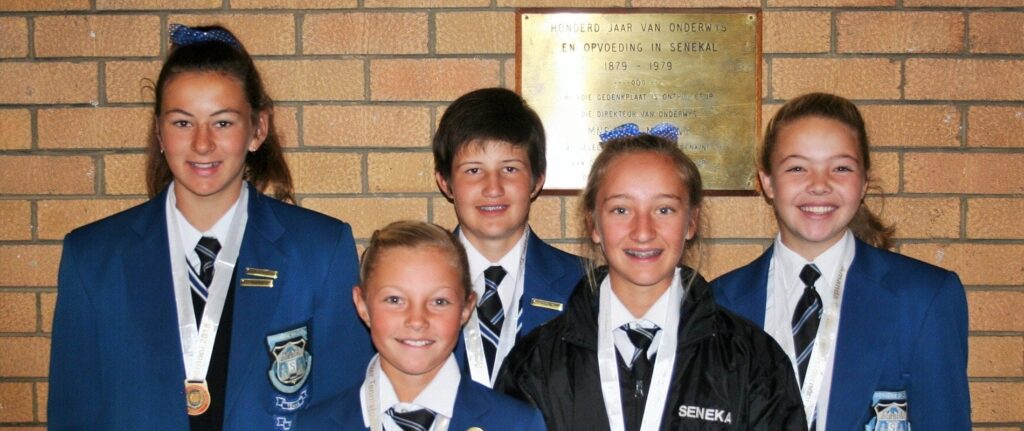 School students wearing blue uniforms and medals, standing against a brick wall with a commemorative plaque, symbolise academic achievement and school ratings in South Africa.